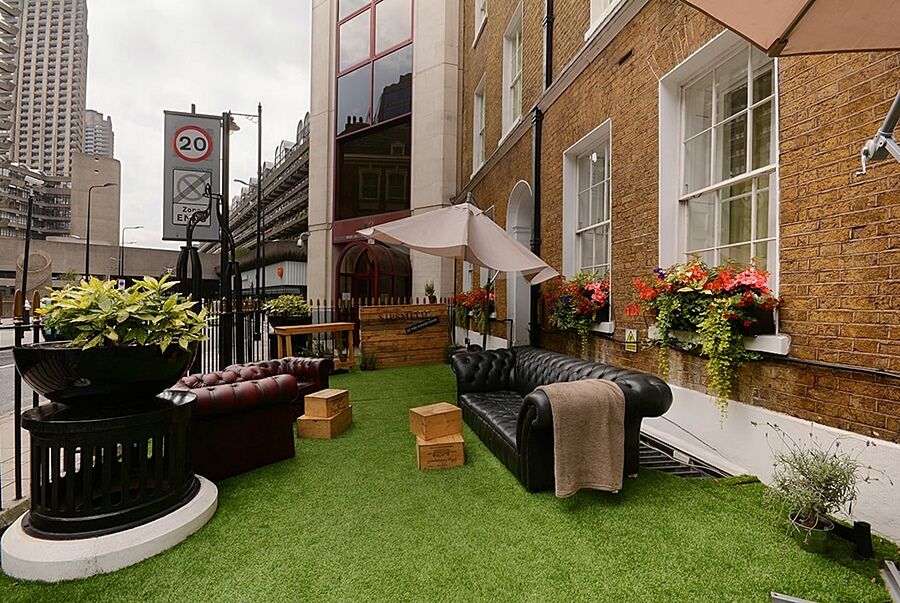 Montcalm Brewery Townhouse Ξενοδοχείο Λονδίνο Εξωτερικό φωτογραφία The photo depicts a charming outdoor space, likely a patio or garden area. It features two comfortable-looking leather sofas, one of which is a deep burgundy color, placed on a green artificial lawn. There are wooden crates that could serve as side t